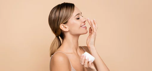 Muurstickers joyful woman with closed eyes applying face cream on nose isolated on beige, banner. © LIGHTFIELD STUDIOS