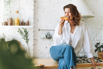 Gorgeous young woman plus size body positive in blue jeans and white shirt eating pizza in the home...