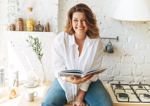 Gorgeous happy young woman plus size body positive in blue jeans and white shirt reading cooking book in the home kitchen