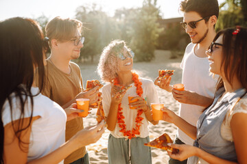 Group of young friends eating pizza, toasting with beerus and have fun on the beach. Happy friends resting together sitting near the sea. Fast food concept. Summer holidays, vacation, relax.