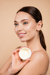 pleased woman holding container with cosmetic cream isolated on beige.