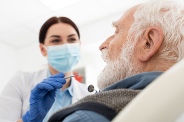 dentist in medical mask examining teeth of senior man with probe and mirror in dental chair.