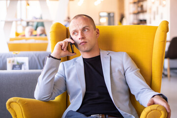 Young business man sitting on a yellow chair in a cafe and phoning. He is arranging a working...