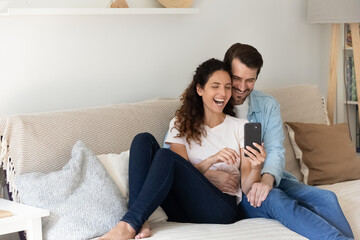 Happy bonding young family couple resting on couch, watching funny photos in social network, having fun entertaining playing mobile games together. Laughing spouses holding web camera video call.