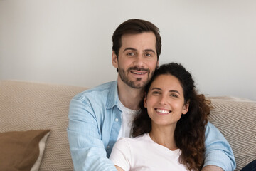Head shot portrait of relaxed millennial generation loving bonding family couple sitting on comfortable sofa. Happy affectionate man cuddling beloved wife, enjoying peaceful moment at own home.