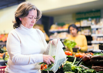 Closeup portrait of elderly woman putting fresh green beans in plastic bag while shopping in...