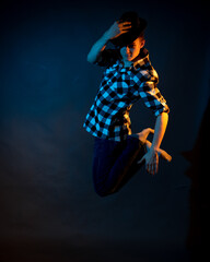 A young man in a plaid shirt jumps on a dark background illuminated by blue and yellow light.