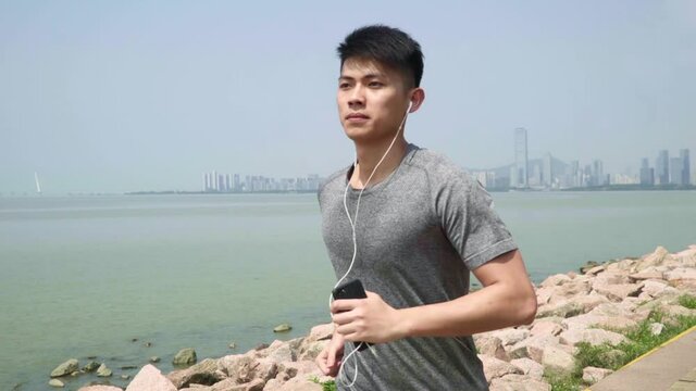 young asian man listening to music while running jogging outdoors on beach by the sea
