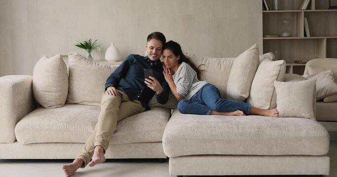 Hispanic couple relaxing on sofa with mobile phone using gadget watching funny videos on internet, making videocall, spend carefree weekend online holds wireless device. Modern tech usage, fun concept