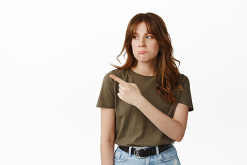 Obraz premium Upset girl show unfair thing, pointing and looking left with sulking sad face, complaining, staring with jealous disappointed expression, standing against white background