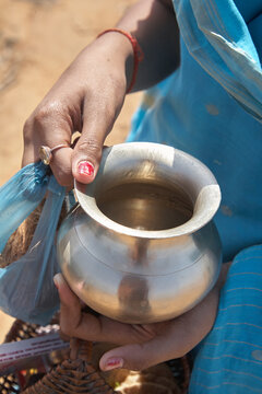 A woman wearing a sari is holding a container (lodha) in her hand and in a plastic bag is a coconut. In the container is probably coconut milk for the puja. India. Day.