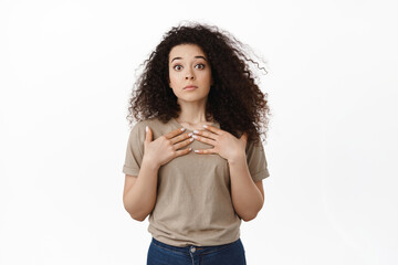 Surprised young woman looking startled, raising eyebrows in disbelief, holding hands on chest, being chosen unexpectedly, staring confused at camera, white background