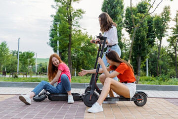 Three Young girl friends on vacation having fun driving electric scooter through the city park. Ecological and urban city transport, summer leisure activities concept