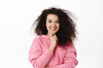 Image of confident brunette woman touch chin, smiling and looking determined at camera, standing in hoodie against white background, have interesting idea