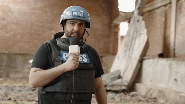 War journalist correspondent wearing bulletproof vest and helmet reporting live near destroyed building