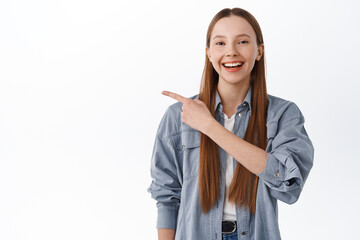 Cheerful college girl with long hair pointing aside, smiling and showing logo, this way gesture,...