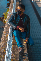 Fashion lifestyle, portrait of a young Latino in the river of the city. Jeans, leather jacket and brown shoes. In a pandemic with a mask