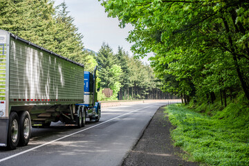 Big rig blue classic semi truck transporting cargo in covered low profile bulk semi trailer driving on the turning road with forest on the sides