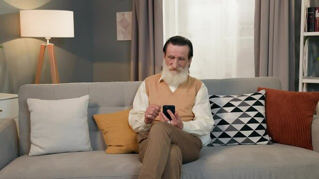 Old Man Sitting On Sofa With Phone Hands. He Has Gray Beard Mustache And Wrinkles On His Face. He Talks To Someone On The Phone Over Video And Shakes His Finger.
