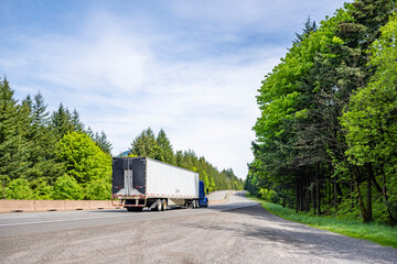 Blue big rig semi truck transporting cargo in dry van semi trailer with aerodynamic tail on the back running on the curving road in Columbia Gorge