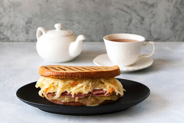 Reuben sandwich is a classic American hot sandwich with sliced beef, Swiss cheese, sauerkraut and sauce. Teapot and a cup of tea on the table.