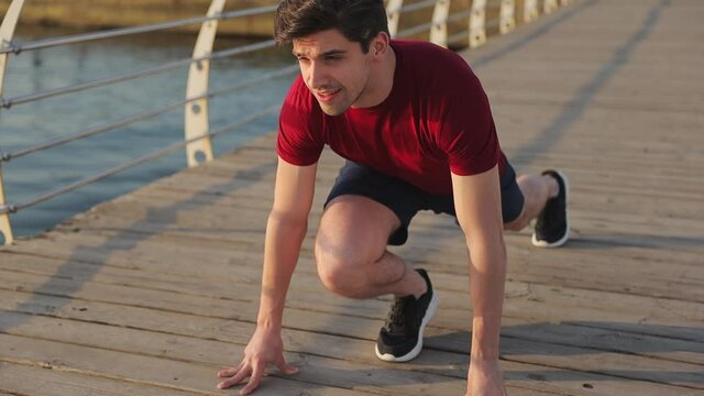 Young strong sporty athletic sportsman man 20s wear red t-shirt black shorts warm up training stand at low start going to run at sunrise over sea beach outdoor jog on wooden pier seaside in summer day