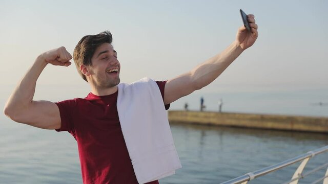 Handsome strong sporty athletic sportsman man 20s wear red t-shirt black shorts warm up doing selfie shot on mobile cell phone training at sunrise over sea beach outdoor on pier seaside in summer day