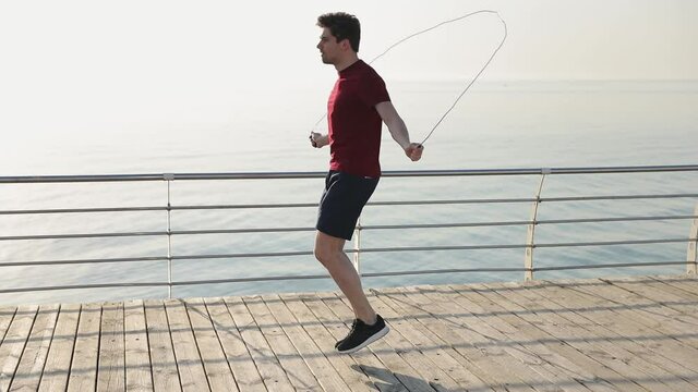 Young strong sporty athletic sportsman man 20s wear red t-shirt black shorts warm up training jump high on skipping rope at sunrise over sea beach ocean outdoor on pier seaside in summer day morning