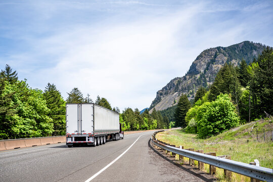 Day cab burgundy big rig semi truck transporting cargo in dry van semi trailer driving on the wide highway road with trees and mountain on background