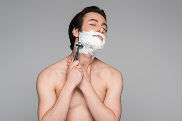shirtless man with white foam on face shaving with safety razor isolated on grey.