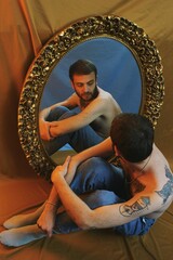 Obraz na płótnie Canvas Studio portrait of young man with tattoos looking at himself in vintage mirror in brown blue background, about self image, gender identity and body positivity 