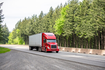 Popular red bonnet big rig semi truck with dry van semi trailer running on the divided highway road with green trees on the side © vit