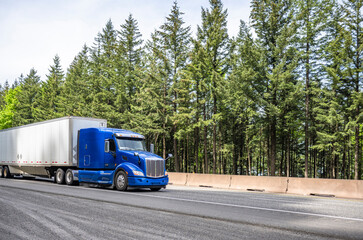 Fototapeta na wymiar Powerful industrial long haul blue big rig semi truck transporting goods in dry van semi trailer running on the wide highway road with trees on the side