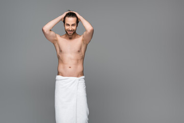 Fototapeta na wymiar happy muscular man wrapped in towel adjusting hair isolated on grey.