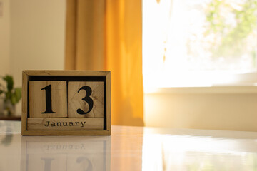 Wooden blocks of the calendar represents the date 13 and the month of January on the background of a window, curtain and a plant.