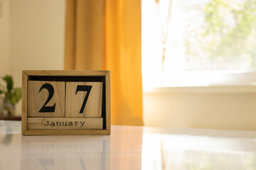Wooden blocks of the calendar represents the date 27 and the month of January on the background of a window, curtain and a plant.