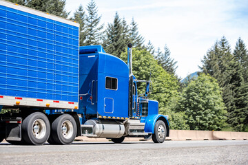 Stylish blue big rig classic bonnet semi truck with refrigerator semi trailer running on the divided highway road with trees on the side