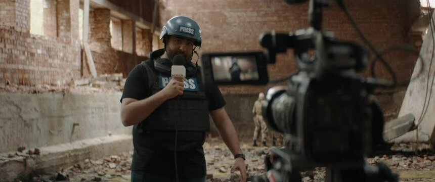 Behind the scenes of war journalist correspondent wearing bulletproof vest and helmet reporting live near destroyed building, military personnel in the background