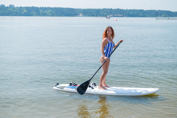 Caucasian woman in a striped swimsuit rides on a SUP board. The girl prefers active rest.