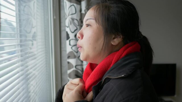 Asian woman wearing black coat and standing at the window, getting cold and looking outside through the window, quarantine and stay safe during Covid 19