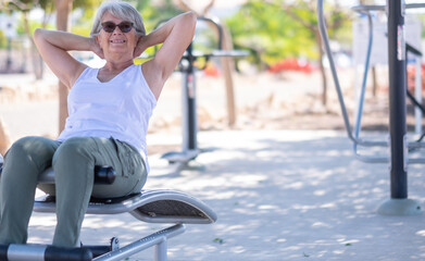 Attractive senior woman in sport activity in public park, sitting on fitness equipment  doing exercises, smiling