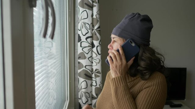 Asian old woman standing at the window and looking outside through the window then taking a call, having a conversation seriously, getting angry. Serious talk on phone. Quarantine concept