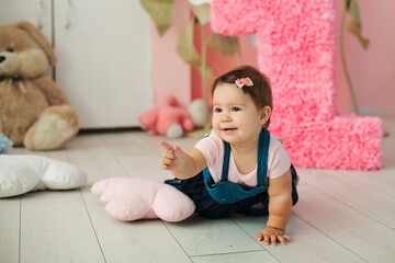 Little cute girl celebrates her birthday at 1 year old. Little girl shows her finger against the background of the number one, fairy flowers and a pink wall