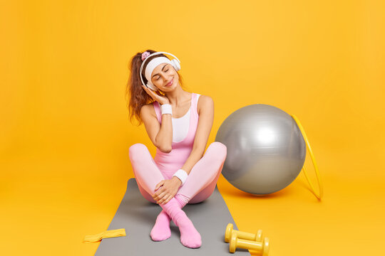 Attractive sporty woman listens audio track feels relaxed and satisfied after fitness training in gym surrounded by sport equipment poses on fitness mat against yellow background. Sport concept