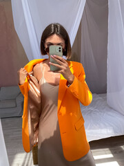 Fit tanned woman in romantic beige silk dress and orange blazer at home take photo selfie on phone...