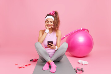 Obraz na płótnie Canvas Fitness sport and training concept. Pleased sporty woman uses smartphone dressed in sportsclothes surrounded by pilates ball headphones does flexibility exercises isolated over pink background