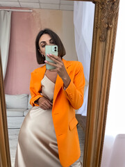Fit tanned woman in romantic beige silk dress and orange blazer at home take photo selfie on phone in mirror for social media, stories, vertical