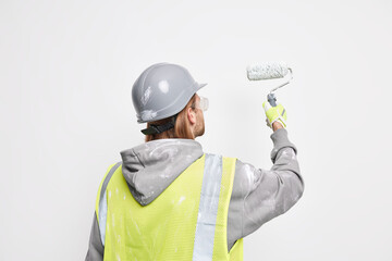 Maintenance service and home improvement concept. Professional male builder stands back at camera wears protective helmet safety uniform paints surface with painting roller follows repair steps