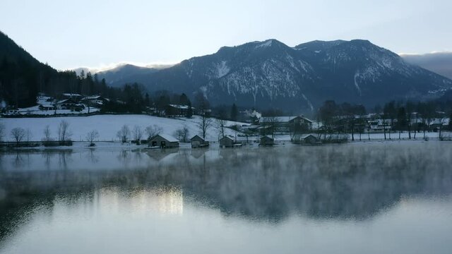 Aerial Panning Lake Shoreline Village Buildings In The Bavarian Alps, With Snow Covered Fields, A Cool Dawn Sky, And Soaring Mountains In The Background - Schliersee, Germany