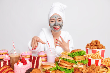 Positive woman applies clay mask for reducing fine lines smiles broadly has good mood dressed in bathrobe poses against different fast food snacks isolated over white background. Harmful nutrition